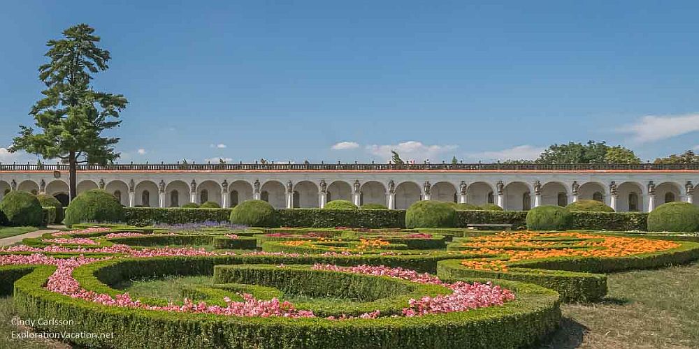 Formal garden in the foreground, with neatly-trimmed hedges surrounding colorful flowers. In the background, a long, white portico edges the garden.