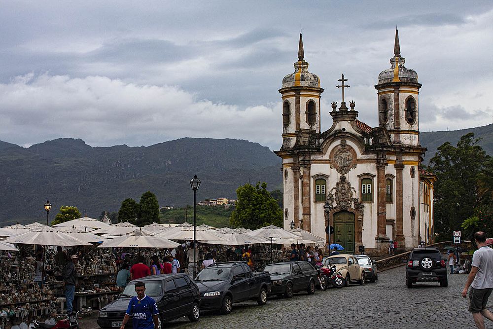 Looking down a street, a row of market stalls along the street with roofs over them. Beyond them, the church: white with brown trim. Two towers, one on either side of an ornate entrance. The church isn't very large: just about 2 stories tall in the front, with an additional story or two in the two towers. 