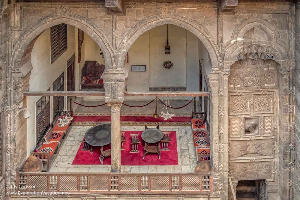 Looking down into a room through huge arched windows without glass. The exterior is delicately carved. In the room are low sofas on the left and right walls, and wooden tables on a rug in the center.