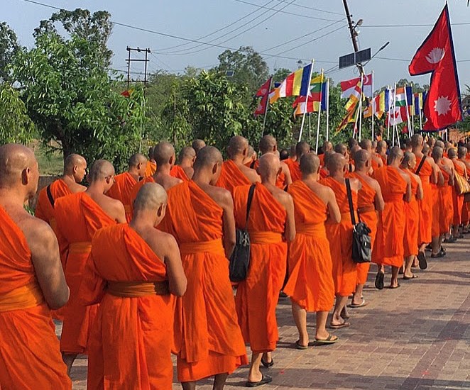 In Lumbini, Nepal, 3 columns of monks in orange robes in a procession, some holding colorful flags, all with shaved heads.