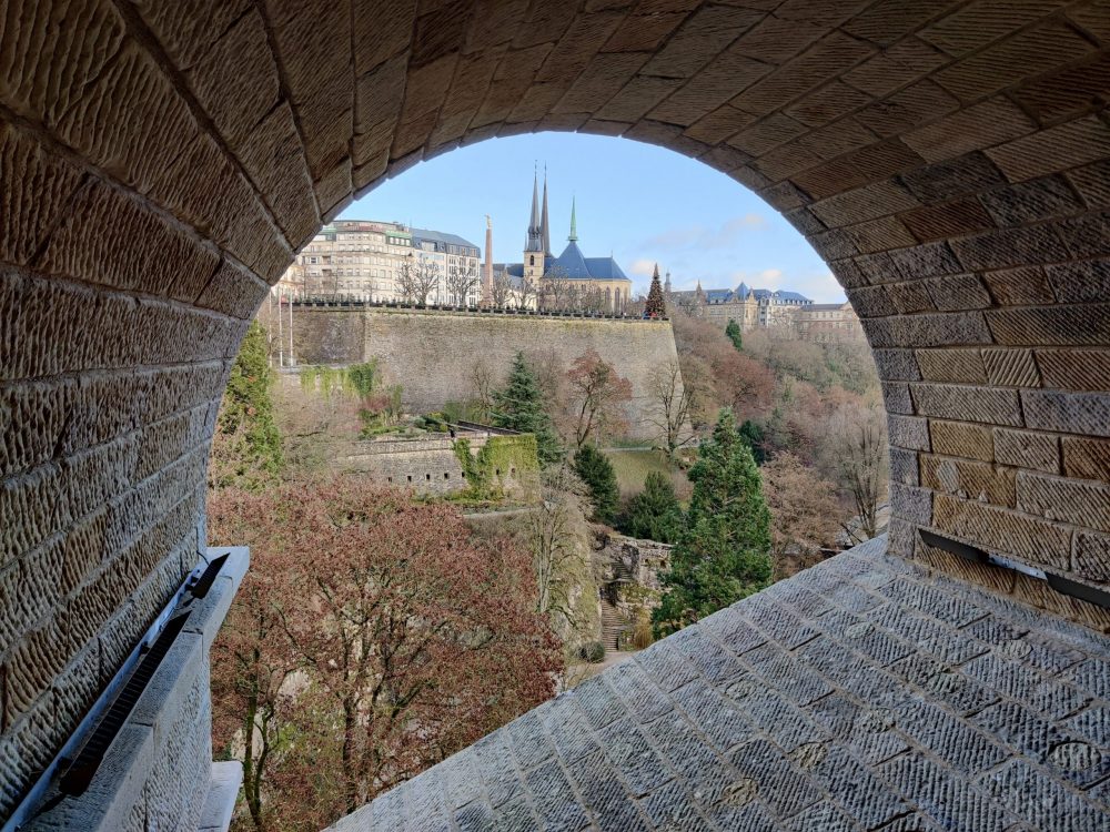 Looking through an archway in Luxembourg City across a valley to the other side, where fortification walls are visible and a church on top and beyond the fortification.