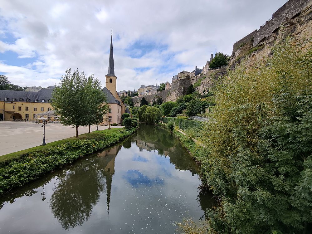 Looking along a small river or canal. On the right, high stone fortifications of Luxembourg City. On the left, a small simple church with a tall spire.