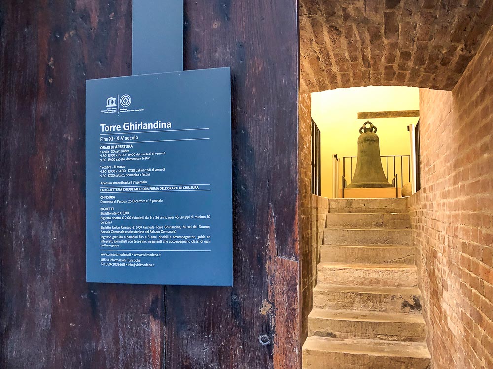 the entrance to the Torre Ghirlandina in Modena, Italy: a sign tells information about opening hours and admission fees. Ahead, a stairway up in a brick passageway, with a bell standing at the top.