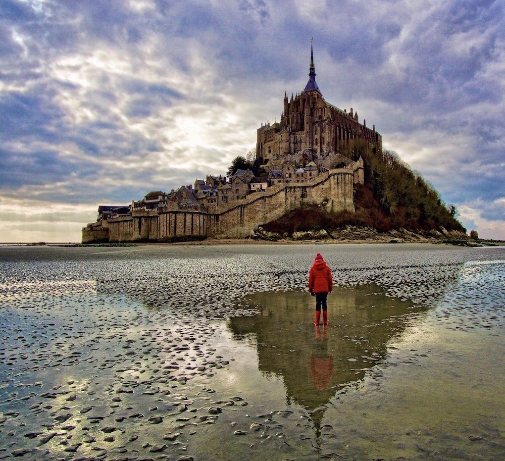 As seen from the sea side of the island at low tide, Mont-Saint-Michel has a fortification wall zig-zagging up the hill, and the Gothic church at the peak. A person stands on the wet mud in the foreground, wearing a red jacket, red had and red rainboots.