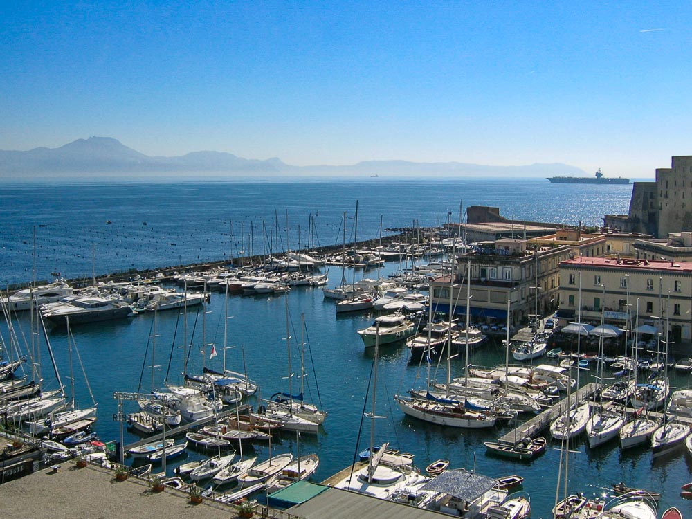 A view over the port of Naples: calm blue water and lots of masted sailboats moored in rows.