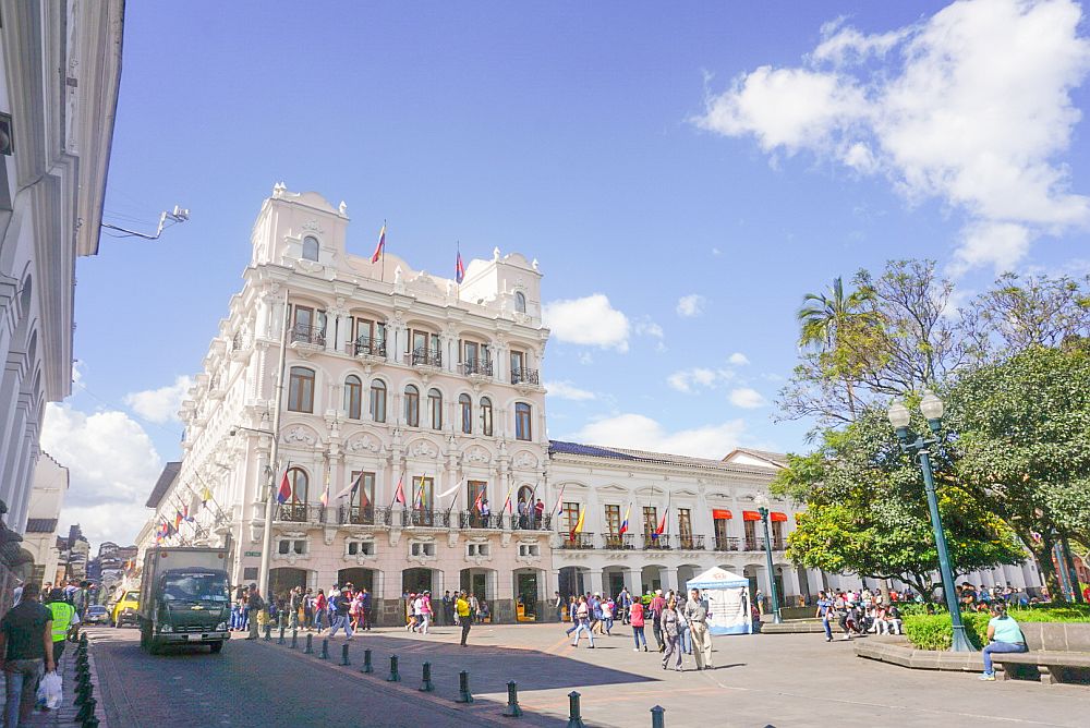 White buildings around a plaza in Quito, Ecuador. They have decorative moldings around windos and doors, and the one in the center of the photo, the corner of the plaza, is higher than most at about 5 stories.