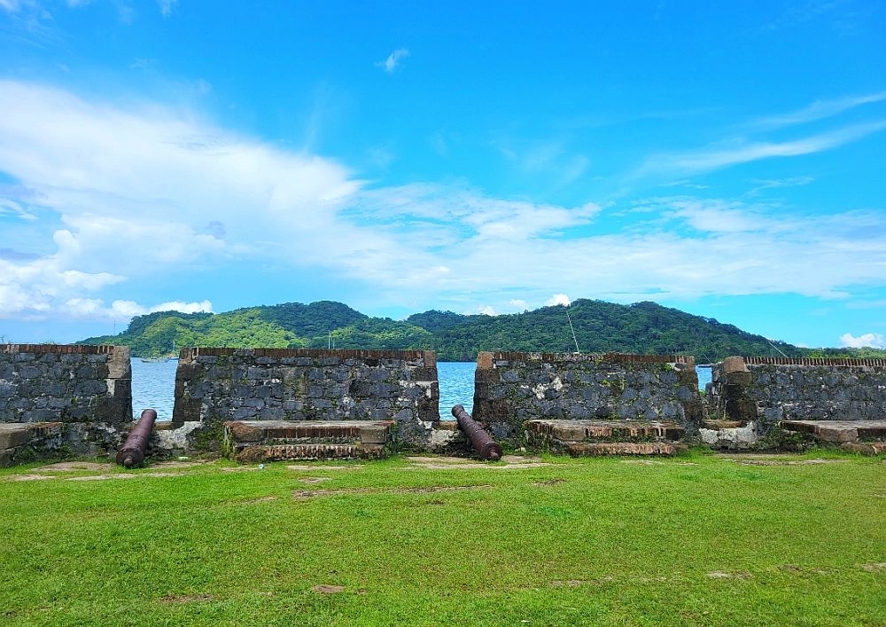 Ramparts in Portobelo town: cannons sit on the ground at each opening in the wall. View over water and wooded land in the distance.