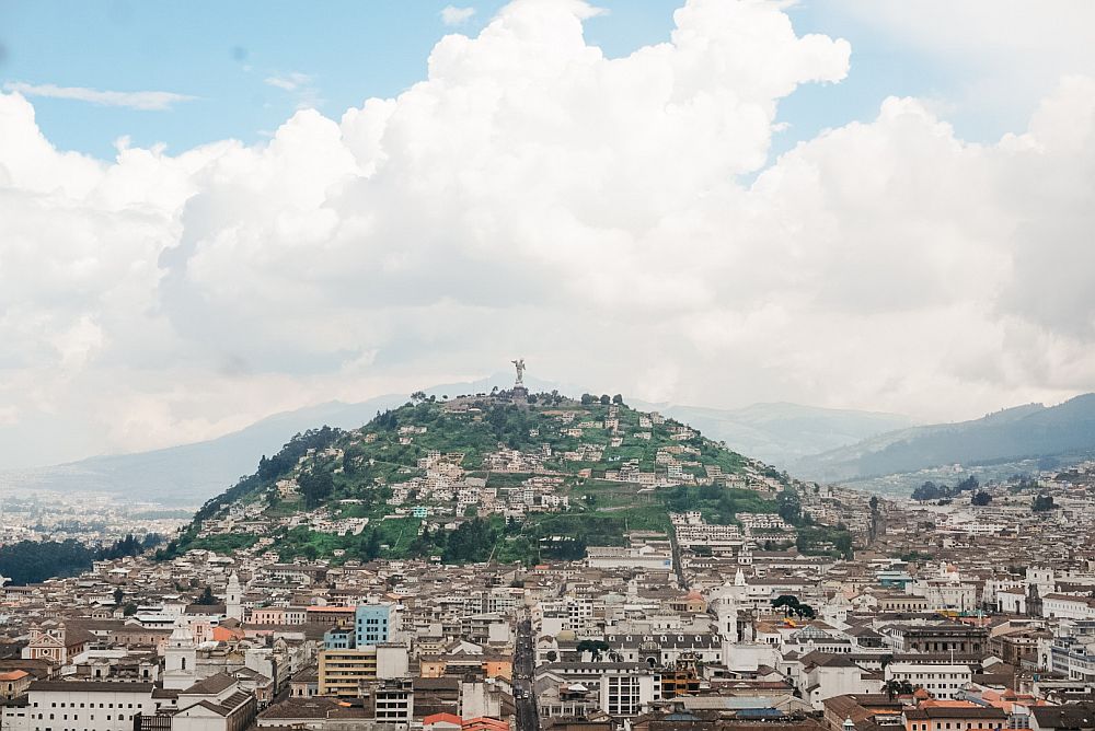 A view of Quito,Ecuador: a flat landscape of blocks filled with buildings. Then, in the background, a bump of a hill with fewer buildings and green between them, and a large statue on the top.