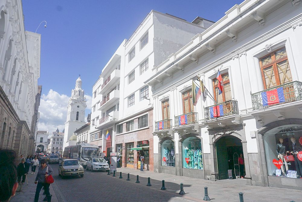 A street in Quito, Ecuador: bright white buildings of varying heights. The nearest has small metal balconies above the ground floor, festooned with flags.
