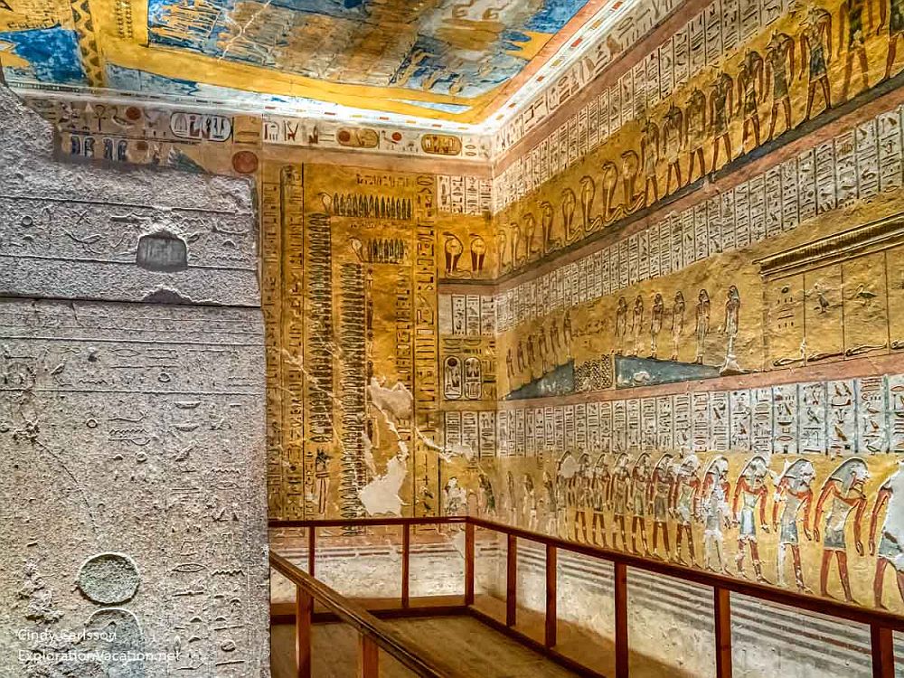 Inside a tomb: the walls and ceiling are entirely filled with paintings and lettering. Rows of people are visible, in the typical Egyptian side view with legs apart. Between the rows of people are very small hieroglyphics filling every space. A small part of a carved tomb is visible on the left, with further hieroglyphics carved into the stone.