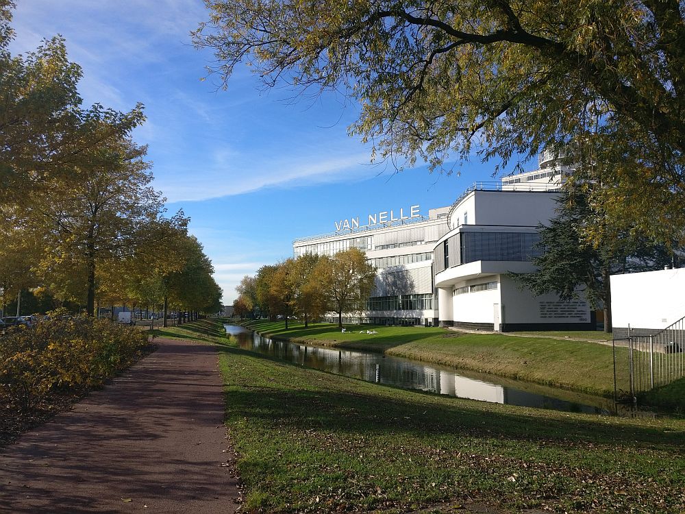 A view of the factory from the side, along a narrow canal. A white building with horizontal lines and rows of windows, and the words Van Nelle standing up on the roof.