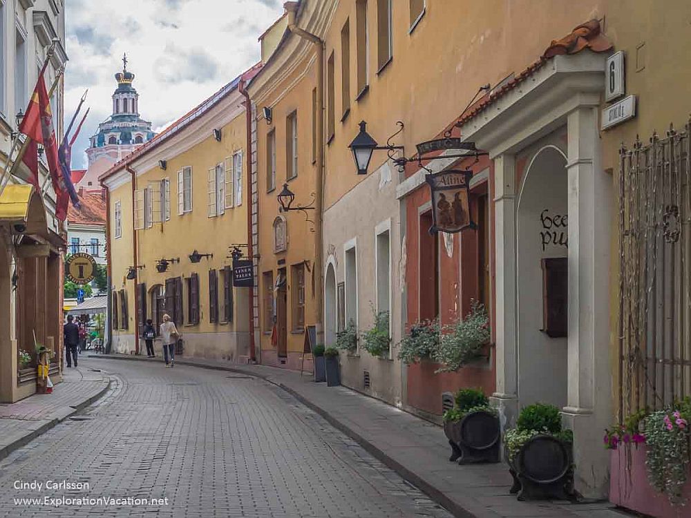 In Vilnius historic center, a street view with what look like medieval era residential buildings along the street, painted in pastels. In the distance a church spire in Baroque style is visible.