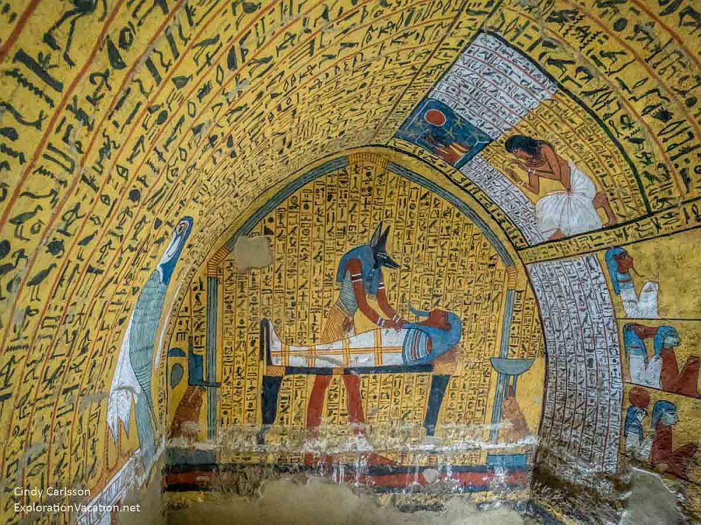 An arched room, walls and ceiling painted with a yellow background. Some paintings are visible, e.g. on the back wall a fox-headed god preparing a mummy on a table, and some smaller images of people on the side walls. Most of the wall and ceiling space around the paintings is filled with hieroglyphics.