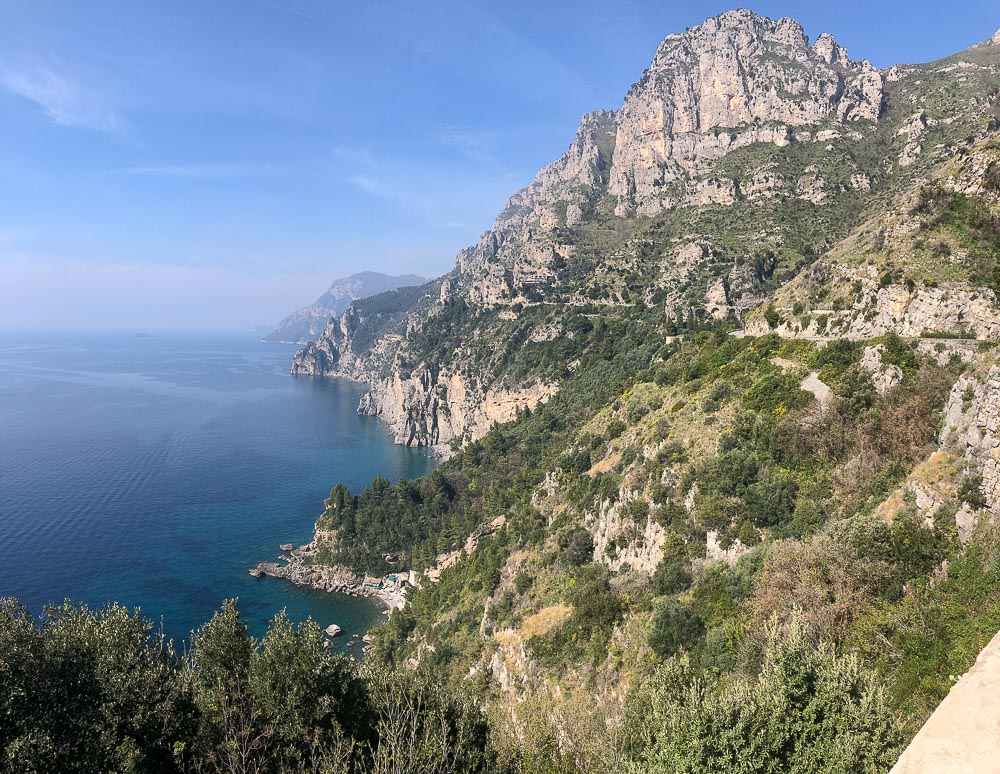 A view along the Amalfi Coast: In this view, no buildings, just jagged mountains that slope steeply down to the sea, with scrubby growth here and there.