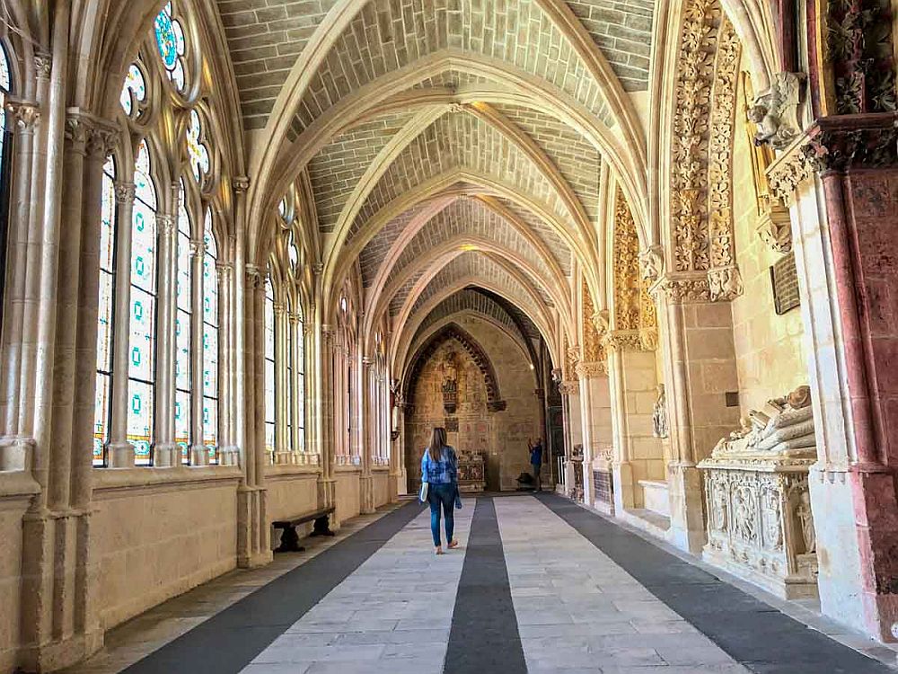 Looking down an aisle or cloister of the cathedral: gothic arches overhead, gothic windows on the left, tombs set between the arches on the right.