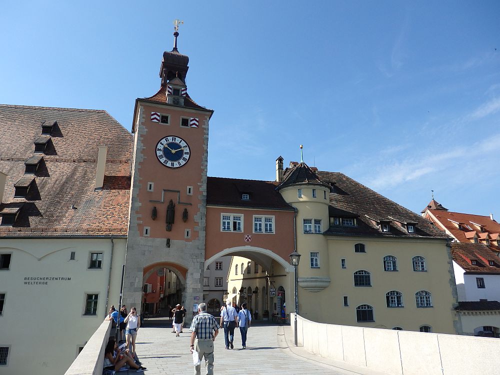 A bridge leads to an archway into Regensburg. Next to the archway, a tall square tower with a large clock.