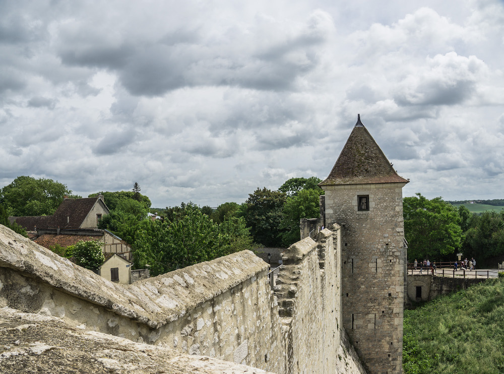A piece of Provins city wall extends down a hill to a round guard tower with a pointed roof.