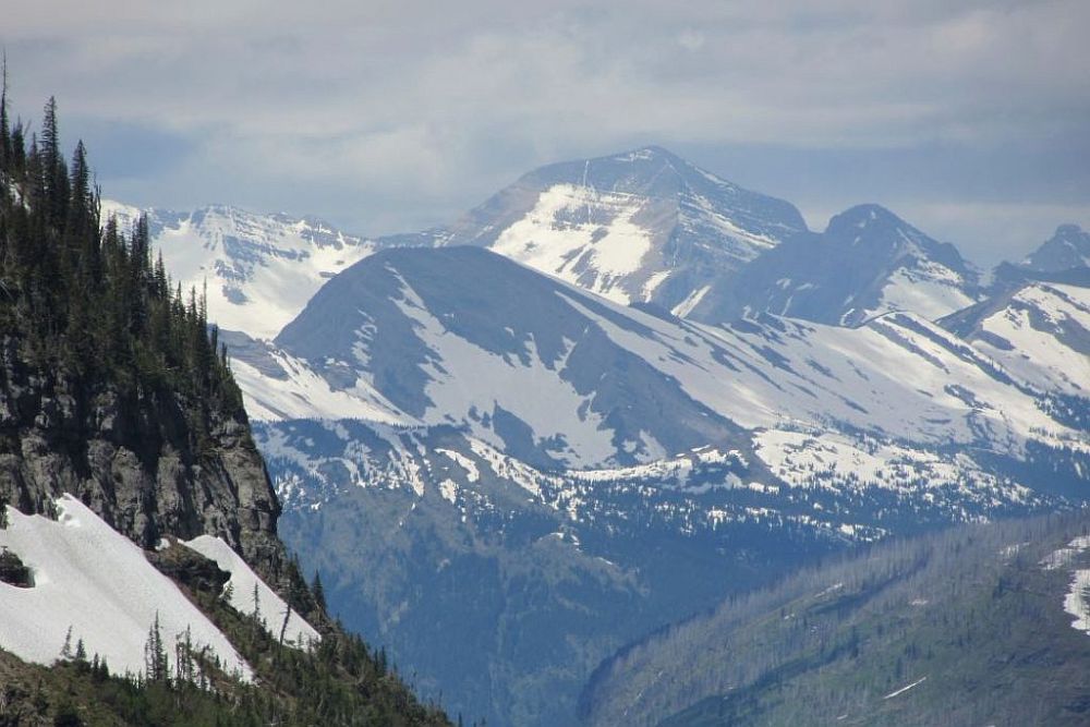 A big view of mountains with snow in the sheltered parts against a cloudy sky in Glacier National Park.