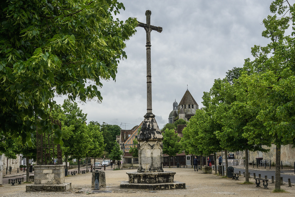 The main square in Provins has a sandy floor and a simple, tall cross in the middle, with rows of trees around the edges of f square. A tower of some kind is partly visible in the background above the trees.