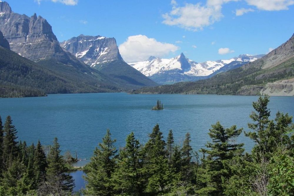 A view over a lake surrounded by rocky mountains topped with snow, pine trees in the foreground and a tiny island in the middle of the lake.