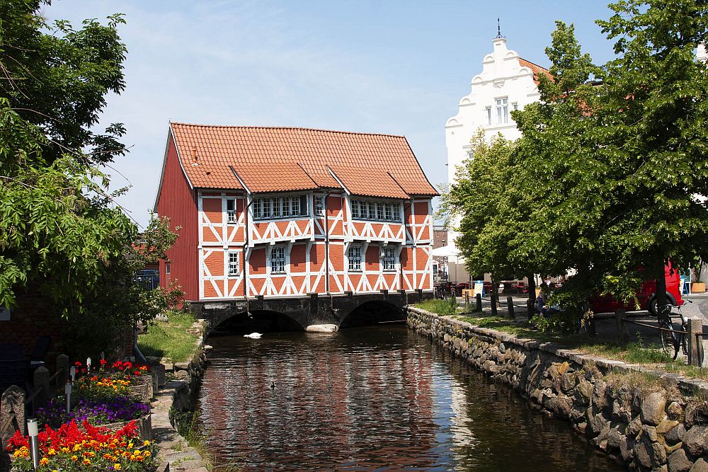 Looking down a canal, a house stands across the canal, presumably on piles in the water. The house is half-timbered, with brick between the beams, which are painted white. The top of a gabled building is partly visible to the right behind some trees.