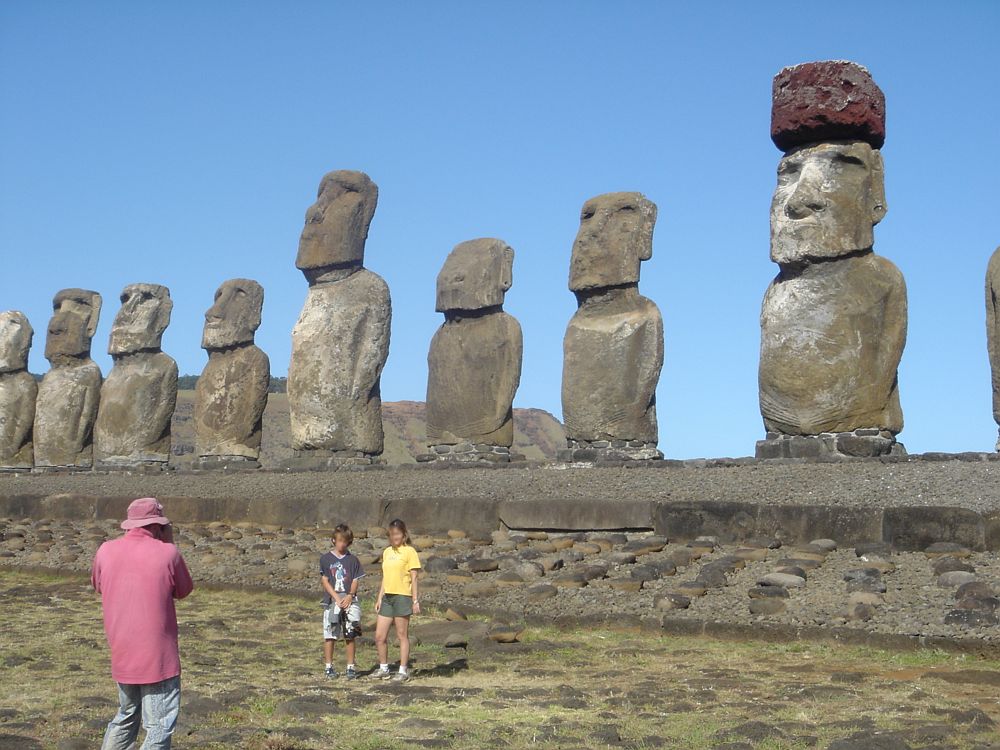 A man takes a photo of his two children posing in front of a row of huge stone figures at Rapa Nui. The bodies are more or less rectangular, and so are the faces. One of them has a large square of reddish stone standing on its head.