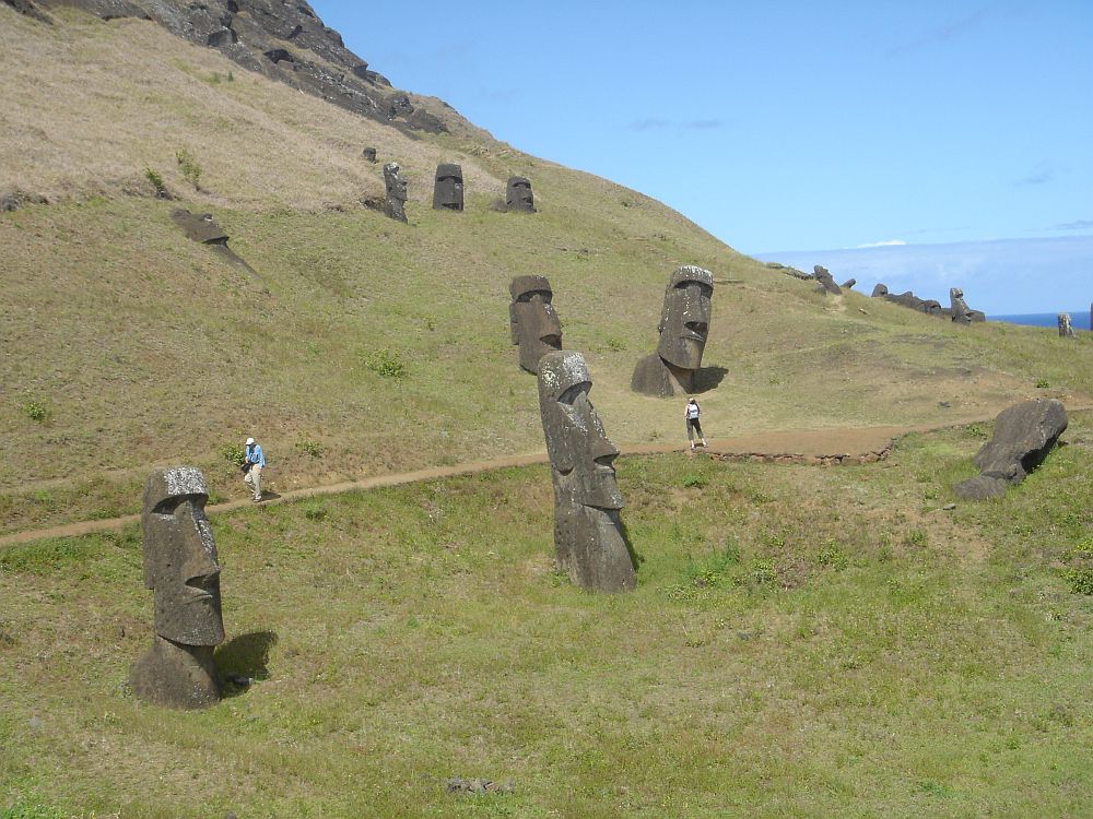 A grass-covered slope at Rapa Nui, with a dirt path crossing it. Huge stone heads stand here and there on the slope, most of them leaning to some extent. A couple of people walk on the path, showing clearly houw huge the figures are: perhaps 3-4x a human's height.
