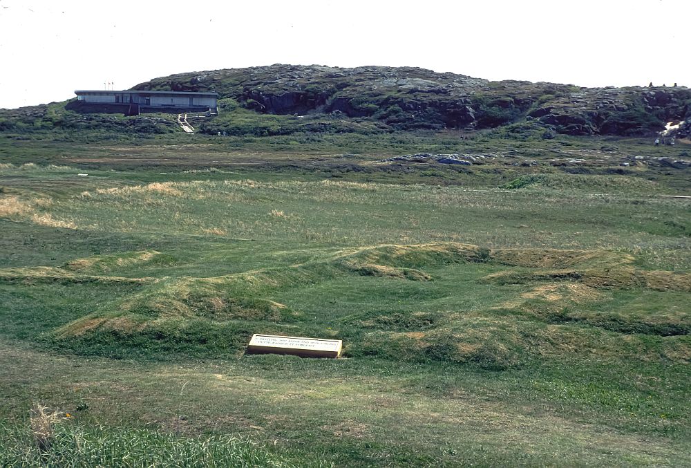 A field with low grass or moss. Low outlines of rectangular building foundations can be seen on the ground. A modern single-story building is in the background.