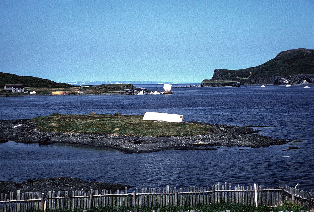 Looking out at the very blue water of the bay at L'Anse aux Meadows, with a Viking ship, with a white square sail in the background.