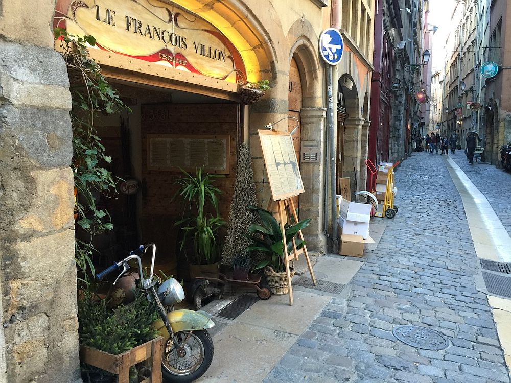 Looking down a cobblestone street, shop entrances with arched stone fronts. The nearest one says "Le Francois Villon" above the shop.