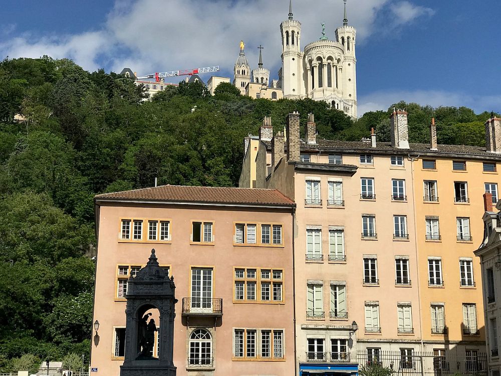 View looking up at Fourviere Basilica on a hill: a white baroque-style confection. At the foot of the hill, a row of older apartment buildings in pastel colors.