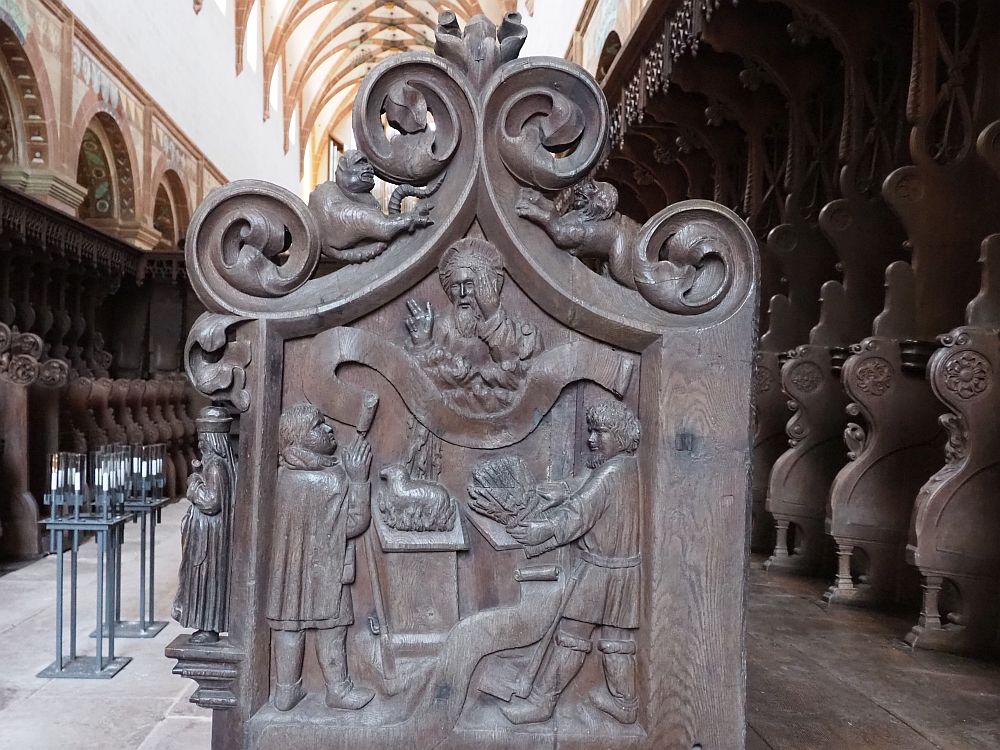A decorative wood panel on the end of the choir stalls in the church: depicts a sacrifice, perhaps, with a lamb on a table, one man to the left praying to a God or Jesus figure above, another man to the right holding some sort of leaves in his hands.
