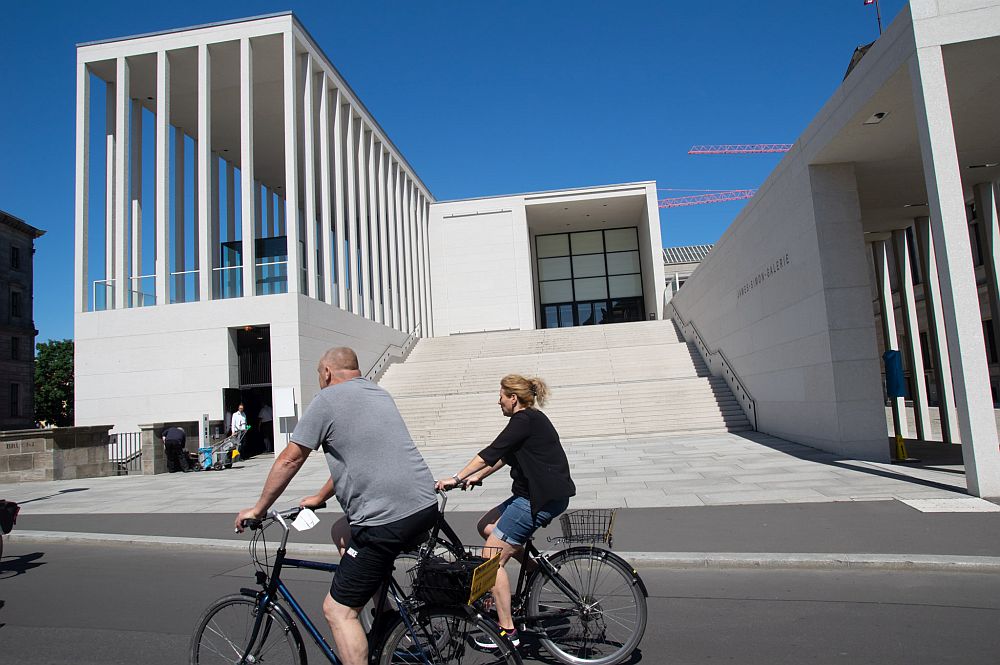 Two people cycle past a very modern white building with a set-back entrance, long stairway leading up to it.