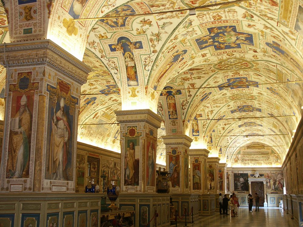 Looking down a long hall in the Vatican that is split lengthwise by a row of pillars. The pillars, the walls,  and the entire arched roof are painted with images of saints and other symbols and images.