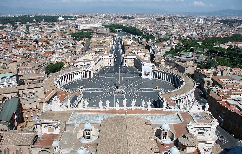 Below where this photo was shot is a line of large statues of saints along the edge of the church's roof. Beyond that is Saint Peter's square, which is round, with pillared colonnades around the sides, except the far section which is open to a straight boulevard. The city of Rome spreads very far in the distance.