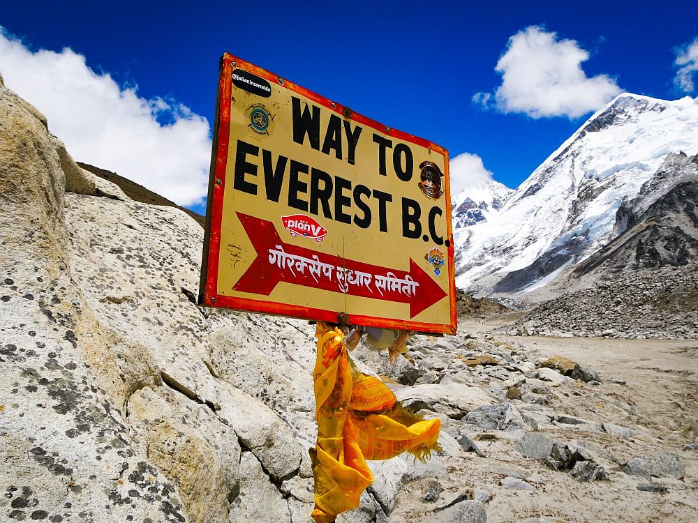 A signpost reads "Way to Everest B.C." with an arrow that has Nepali writing on it. B.C. means base camp. Behind the sign is a mountain covered in snow.