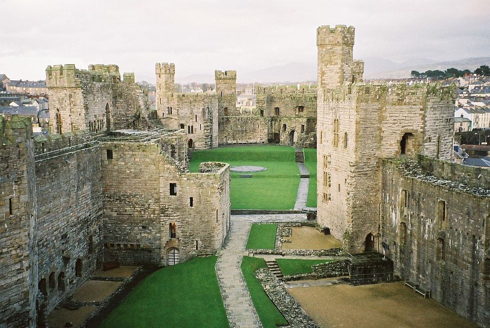 Walls around the side of an irregular grassy courtyard are made of whitish stone, with crenellations around the top, and towers on along the wall's extent. The various towers appear intact, but the rampart seem damaged on the nearer parts of the walls.