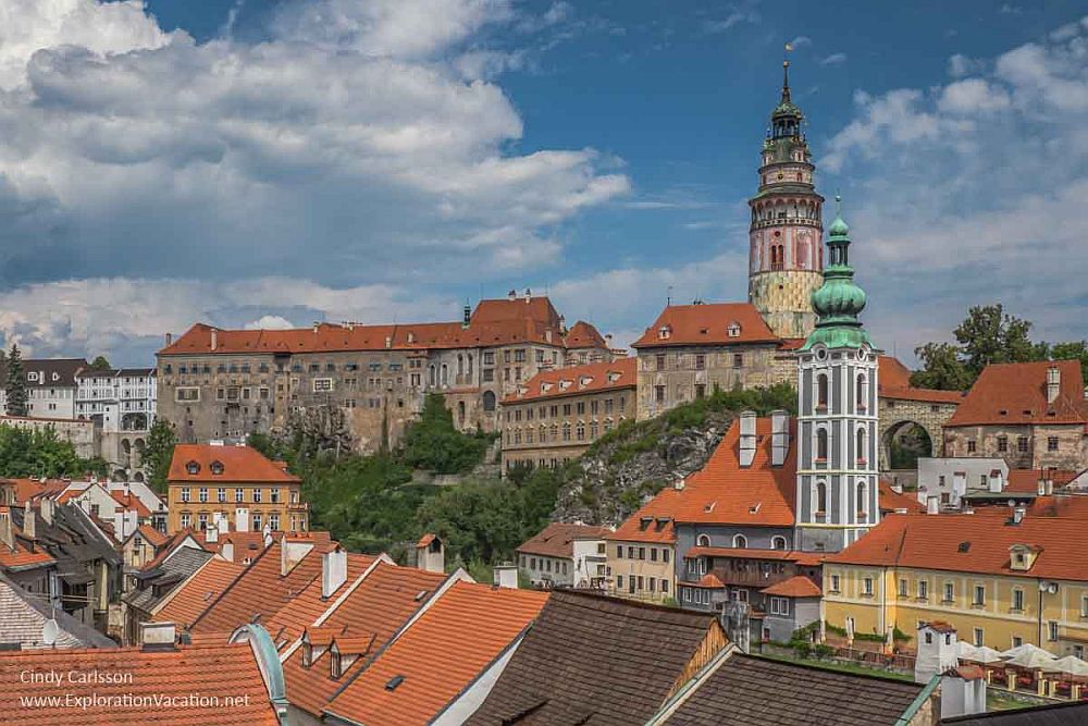A view of Český Krumlov Castle with the red roofs of the town in front of it. The castle is very large and has a round tower rising quite high above it.
