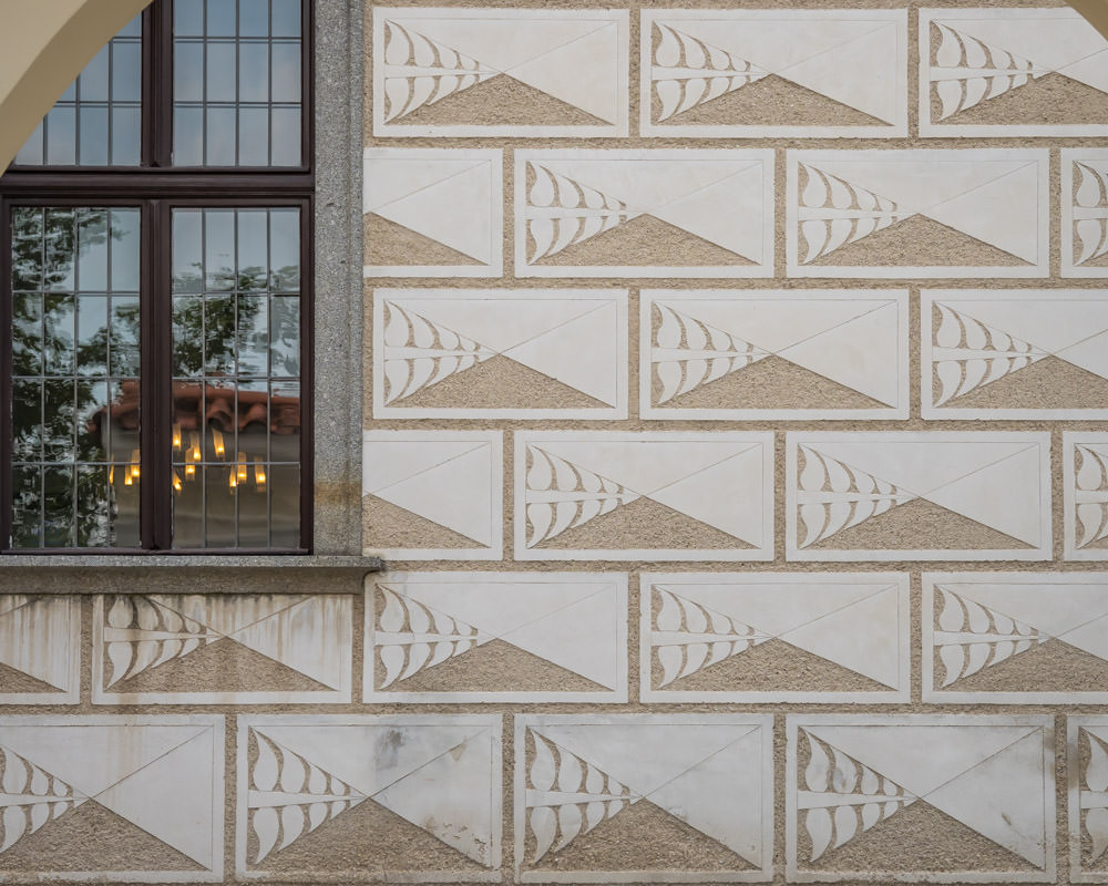 Close-up of a sgraffito patter on a building in Český Krumlov. Bricks are outlined in horizontal rectangles and each brick is divided in 4 parts with a wide x. The bottom space of the x is carved out completely so it makes a brown triangle. The left-hand part of the x has a pattern carved into it - the same on every "brick." The other two parts are left white. 