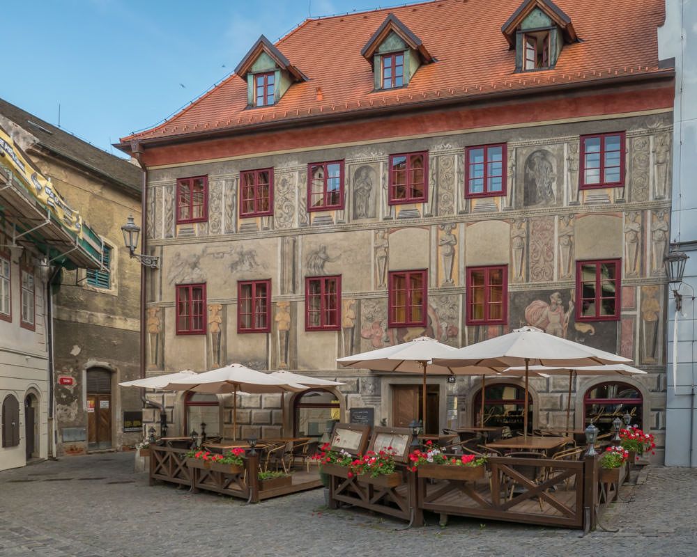 A 4-story house in Český Krumlov houses a cafe - outdoor tables in front with umbrellas over them. The house is painted with trompe-l'oeil images that look like statues carved into niches between the windows.
