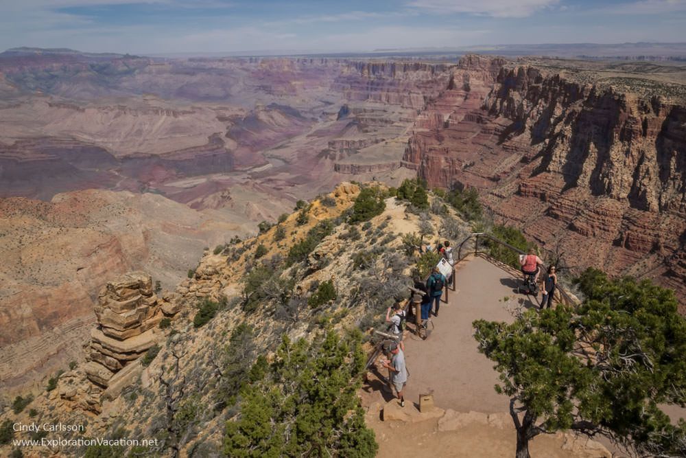 Looking down onto a viewpoint over the Grand Canyon, with people standing along a fence edging an outcropping of rock. Steep red rock cliffs on the right, with horizontal layers of rock visible. On the left, a wider and further view of the canyon.