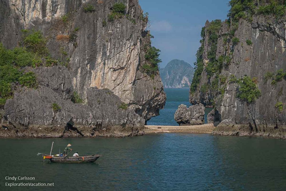 A small fishing boat in Ha Long Bay, in front of two islands connected by a small stretch of sand. Between them more sea is visible.