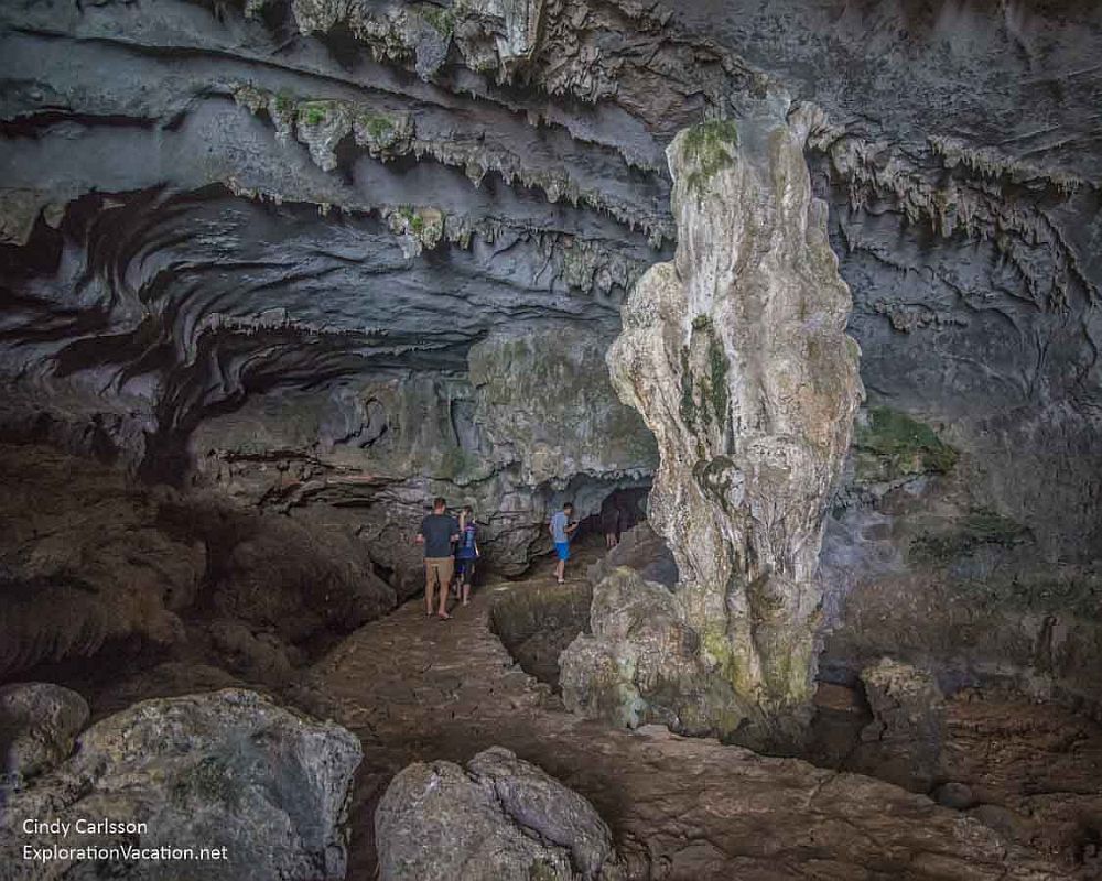 A large cave opening at Ha Long Bay, short stalagmites hanging from the roof, one large natural pillar. A stone path leads deeper into it, and 3 people walk into the opening.