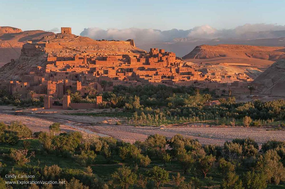 A view of the whole Ksar of Ait-Ben-Haddou, red in the sunrise. It's a jumble of square and rectangular buildings of varying heights, clustered up the side of a hill with a fortress above it.