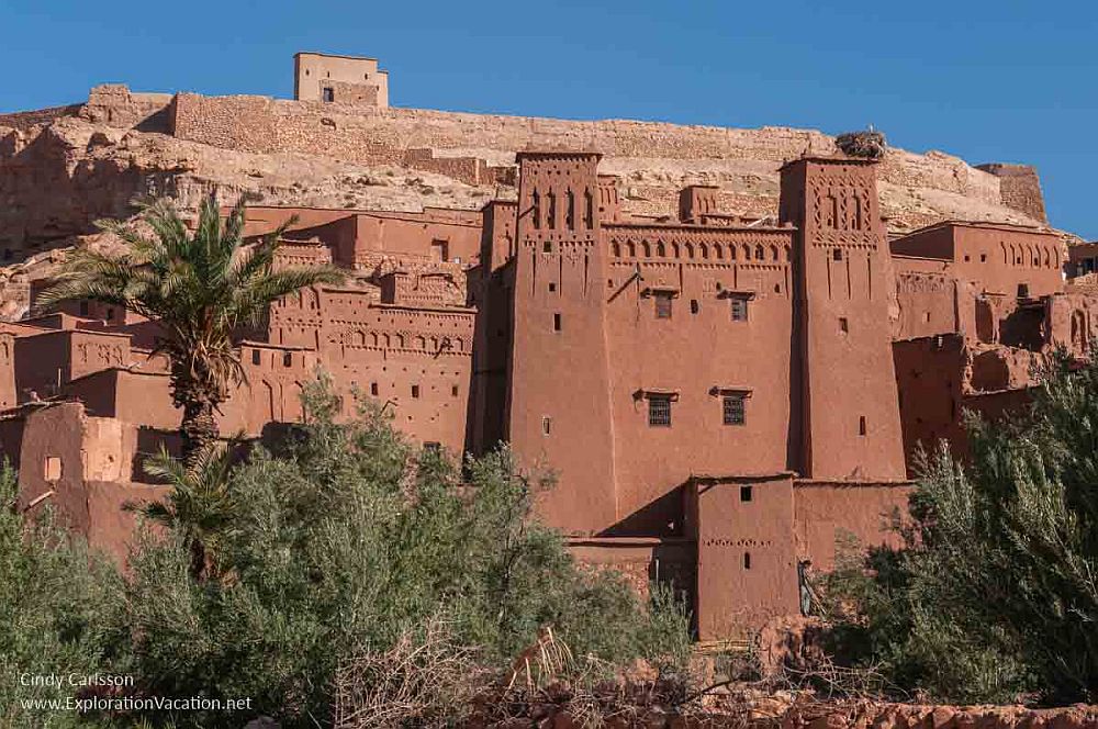 A cluster of red earth buildings with few windows, on the side of a hill. Decorative features carved around the tops of the buildings. Above them, on top of the hill, stone walls of a fortress.