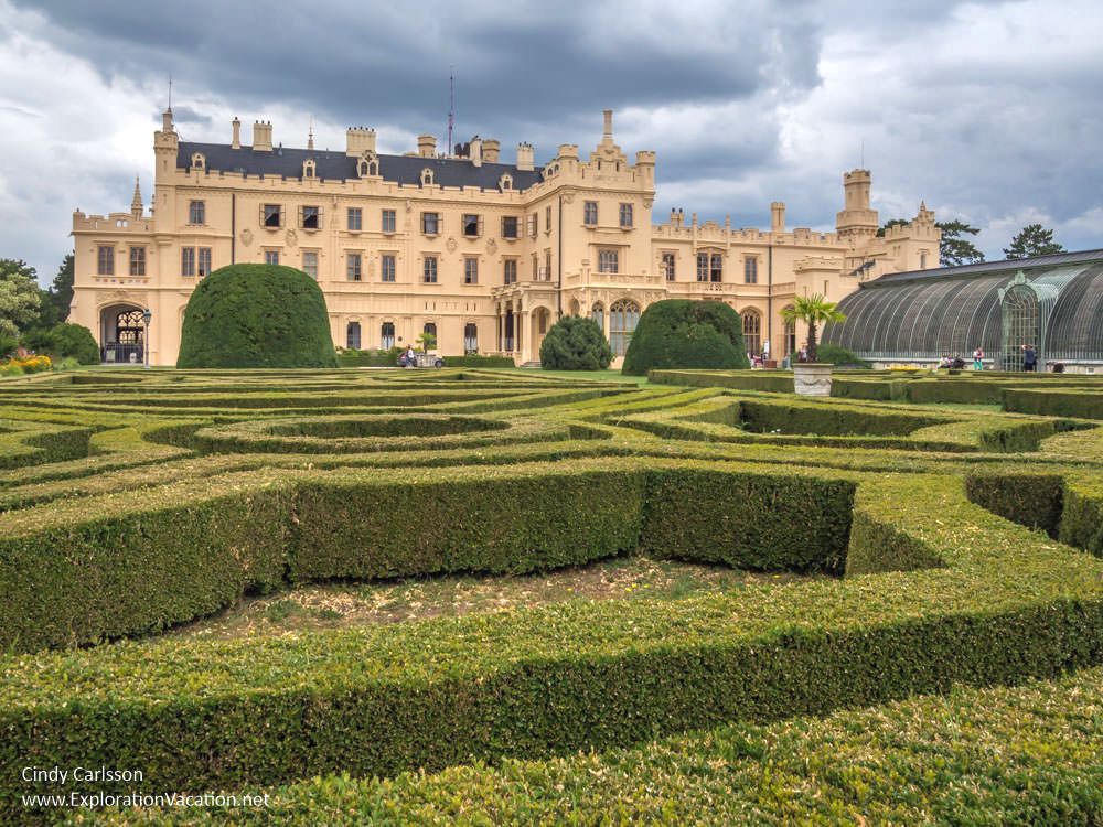Foreground: exactly trimmed hedges outline the walkways in a formal garden. Background: Lednice Castle is 3-4 stories tall and very wide, with some decorative elements along the roof line and around the ground floor.