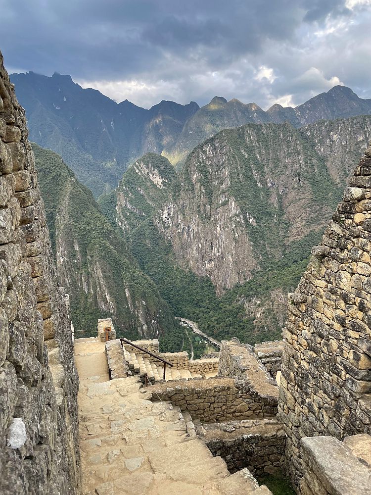 Looking down a stone stairway (no handrail) at Machu Picchu. High stone walls on either side and a view over steep craggy mountains.