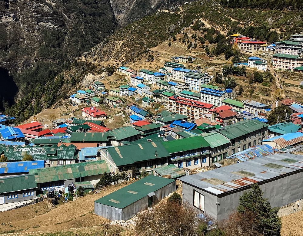 A cluster of houses and apartment blocks on terraces on the side of a hill. Many have either blue or green roofs.