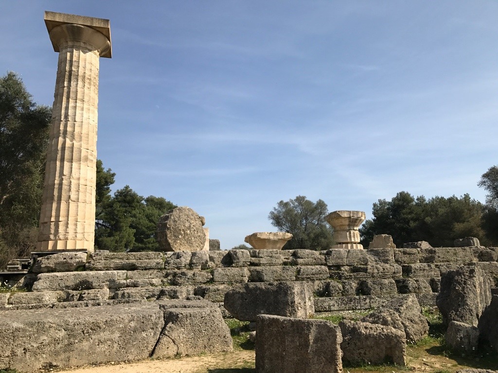 A small part of the ruins: one complete column in marble and several broken ones, in a line on a wall of darker stone at Olympia.