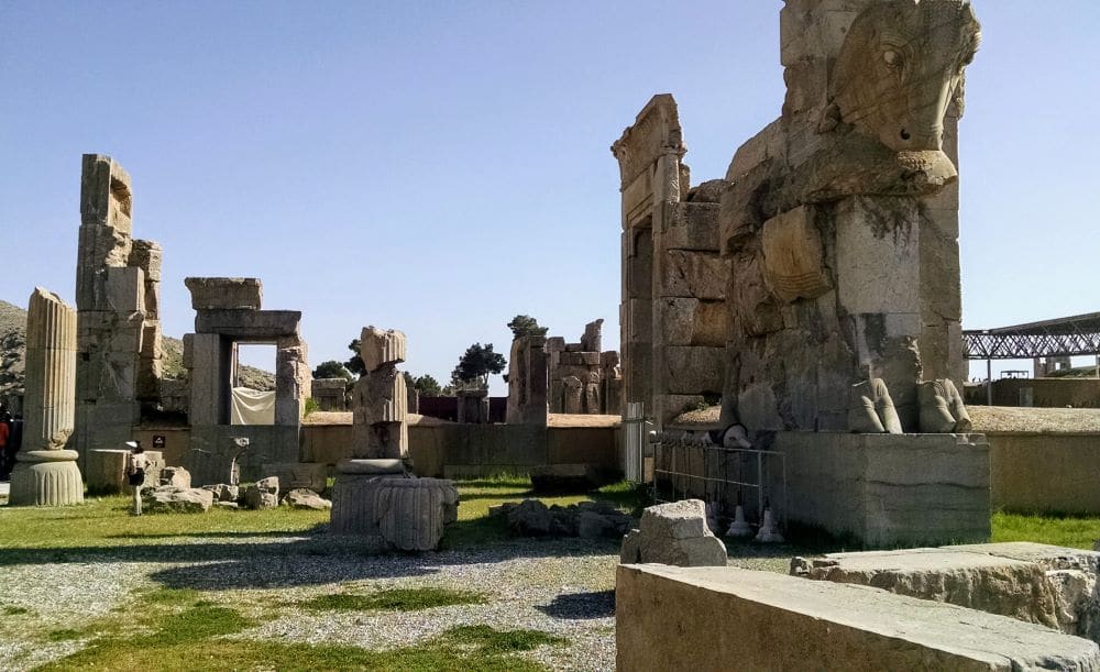 Assorted pieces of the ruins at Persepolis. To the right, a very large stone sculpture of what appears to be a horse or ox. Behind, various broken pillars, and a gateway.